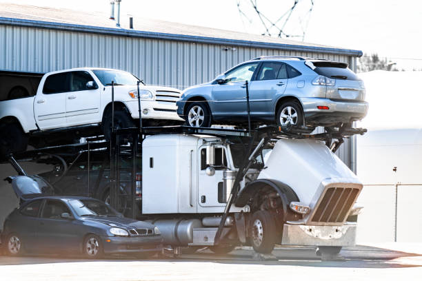A white car carrier truck, part of a Car Transport Portland, OR service, transports three vehicles—two SUVs and a sedan—outside an industrial building in daylight. The truck’s cab is tilted forward for maintenance.